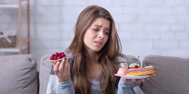 Depressed young lady having eating disorder, comforting herself with sweets on sofa at home. Unhappy millennial woman coping with negative emotions through food, suffering from bulimia